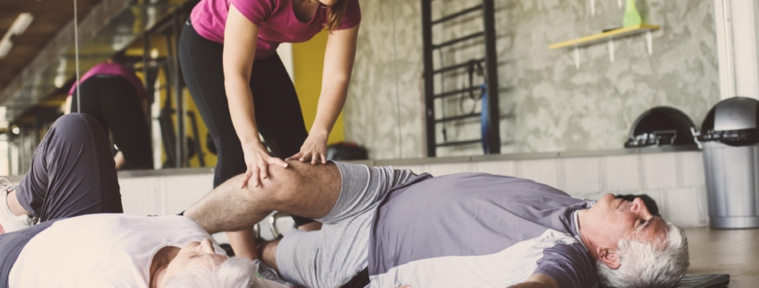 Senior couple workout in rehabilitation center. Personal trainer helps elderly couple to do stretching on the floor.