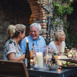 A group of mature friends are sitting around an outdoor dining table, eating and drinking. They are all talking happily and enjoying each others company. The image has been taken in Tuscany, Italy.