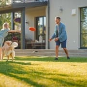 Smiling Beautiful Family of Four Play Fetch flying disc with Happy Golden Retriever Dog on the Backyard Lawn. Idyllic Family Has Fun with Loyal Pedigree Dog Outdoors in Summer House Backyard