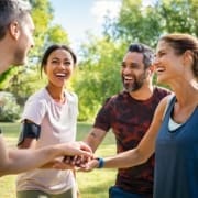 Laughing mature and multiethnic sports people putting hands together at park. Happy group of men and beautiful women smiling and stacking hands outdoor after fitness training. Multiethnic sweaty team cheering after intense training.