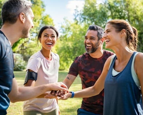Laughing mature and multiethnic sports people putting hands together at park. Happy group of men and beautiful women smiling and stacking hands outdoor after fitness training. Multiethnic sweaty team cheering after intense training.