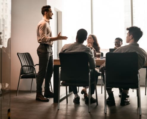 Business Presentation. Smiling Businessman Giving Speech During Seminar With Coworkers In Office, Standing At Desk In Boardroom, Diverse People Sitting At Table And Listening To Speaker