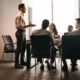 Business Presentation. Smiling Businessman Giving Speech During Seminar With Coworkers In Office, Standing At Desk In Boardroom, Diverse People Sitting At Table And Listening To Speaker