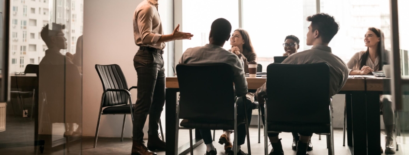 Business Presentation. Smiling Businessman Giving Speech During Seminar With Coworkers In Office, Standing At Desk In Boardroom, Diverse People Sitting At Table And Listening To Speaker