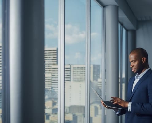 Thoughtful Black Businessman in a Tailored Suit Using Laptop while Standing in Office Near Window on Big City. Successful Corporate Top Manager Doing Data Analysis for e-Commerce Startup