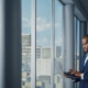 Thoughtful Black Businessman in a Tailored Suit Using Laptop while Standing in Office Near Window on Big City. Successful Corporate Top Manager Doing Data Analysis for e-Commerce Startup