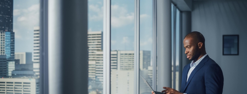 Thoughtful Black Businessman in a Tailored Suit Using Laptop while Standing in Office Near Window on Big City. Successful Corporate Top Manager Doing Data Analysis for e-Commerce Startup