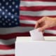 Close-up of man casting and inserting a vote and choosing and making a decision what he wants in polling box with United States flag blended in background..