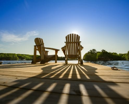 Cottage life - Sunrise on two empty Adirondack chairs sitting on a dock on a lake in Muskoka, Ontario Canada. The sun rays create long shadows on the wooden pier.