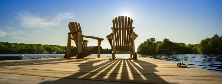 Cottage life - Sunrise on two empty Adirondack chairs sitting on a dock on a lake in Muskoka, Ontario Canada. The sun rays create long shadows on the wooden pier.