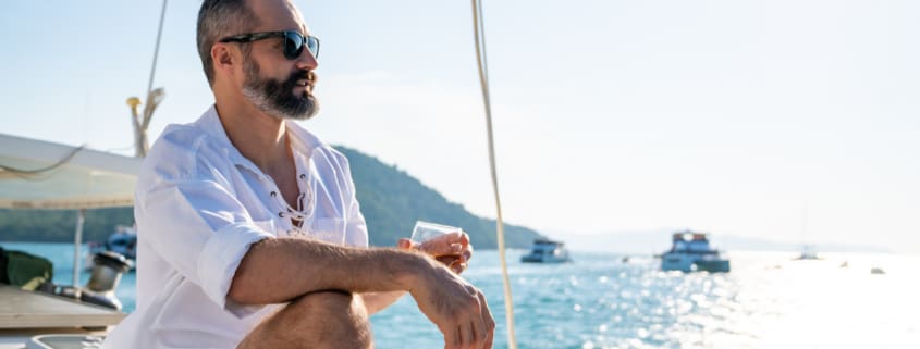 Caucasian man enjoy outdoor luxury lifestyle with alcoholic drinks while catamaran boat sailing at summer sunset. Healthy handsome male relaxing outdoor leisure activity with tropical travel vacation trip